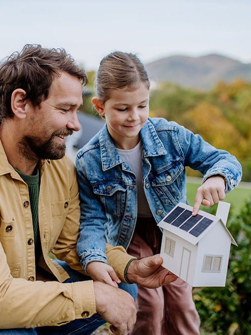 Kleines Mädchen mit ihrem Vater hält ein Papiermodell eines Hauses mit Sonnenkollektoren in der Hand und erklärt, wie es funktioniert. Alternative Energie, Ressourcenschonung und nachhaltiges Lebensstilkonzept.
